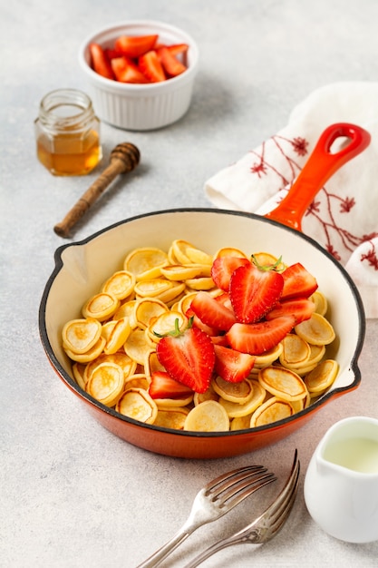 Mini cereal panqueque blanco con fresas en una sartén para el desayuno sobre fondo gris. Desayuno casero de moda con tortitas pequeñas. Vista superior.