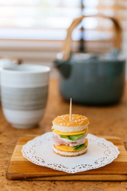 Mini Chicken Burger en la tajadera de madera con la taza y la taza de té de la falta de definición en el fondo.