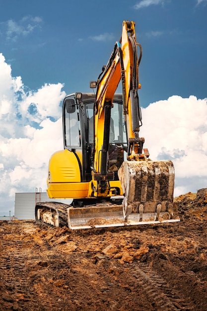 Mini escavadeira no canteiro de obras à beira de um poço contra um céu azul nublado Equipamento de construção compacto para terraplenagem Um assistente indispensável para terraplenagem