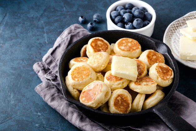 Mini-Getreidepfannkuchen in der gusseisernen Bratpfanne auf grauer Serviette, Blaubeere. Dunkelblauer Zementhintergrund, Draufsicht.