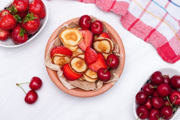 Mini panqueques pequeños con straeberries y cerezas sobre fondo blanco de madera. Concepto de comida de moda. Endecha plana.