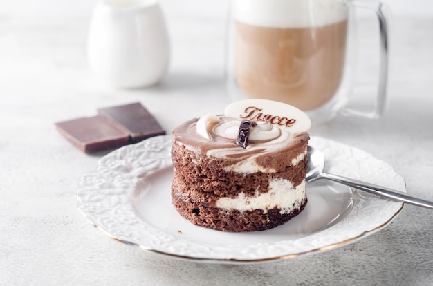 Mini pastel de chocolate en elegante plato blanco y capuchino con espuma en vaso sobre fondo gris claro, vista superior. Postre delicioso. Cubierto de mesa de desayuno.