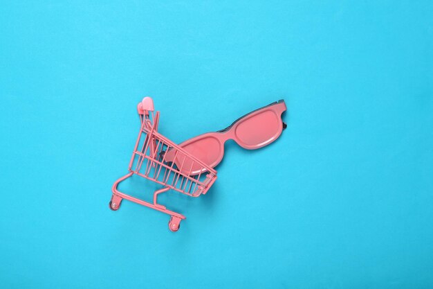 Mini rosa Supermarkt Trolley mit rosa Sonnenbrille auf blauem Hintergrund Ansicht von oben flach legen Konzeptkunst Minimalismus