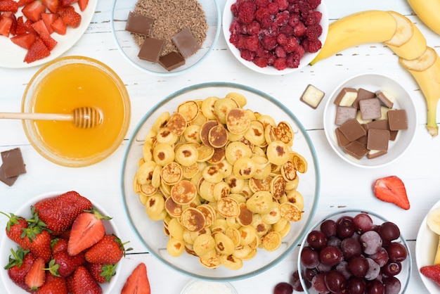 Mini tazón de cereal para panqueques con fresas frescas, chocolate de plátano, virutas de coco y miel en pequeños platos.