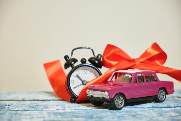 Miniatur lila Retro-Automodell, roter Schleife und Wecker auf Holzoberfläche, grauer Hintergrund
