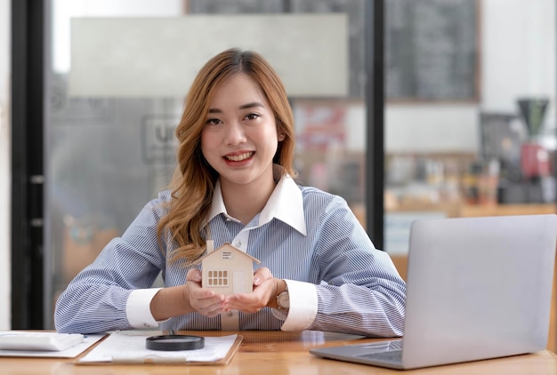 Miniaturhaus in den Händen einer asiatischen Immobilienmaklerin, die im Büro arbeitet und in die Kamera schaut
