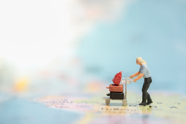 Miniaturleute stellen das Gehen mit Flughafenlaufkatze / -warenkorb mit Gepäck auf Weltkarte dar.