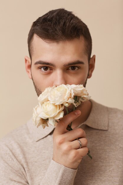 Minimales Nahaufnahmeporträt eines weiblichen jungen Mannes, der Blumen hält und in die Kamera schaut, während er im Studio vor neutralem beigefarbenem Hintergrund posiert