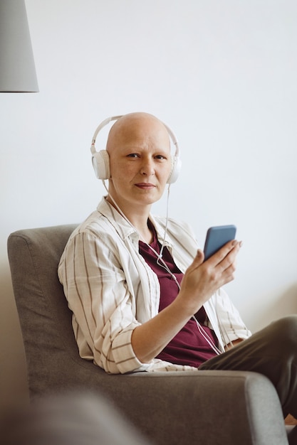 Minimales Porträt der kahlen erwachsenen Frau, die Kamera beim Hören von Musik über Smartphone zu Hause, Alopezie und Krebsbewusstsein, Kopierraum betrachtet