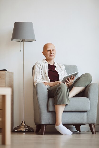 Minimales Porträt in voller Länge der kahlen erwachsenen Frau, die Buch hält und Kamera nachdenklich betrachtet, während im bequemen Sessel zu Hause, Alopezie und Krebsbewusstsein sitzen