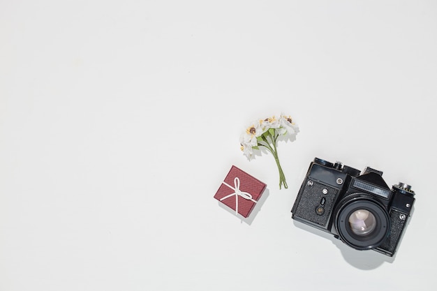 Minimalistische flache Laienkomposition mit Retro-Kamera, weiniger Geschenkbox und Frühlingsfeldblume auf weißem Hintergrund.