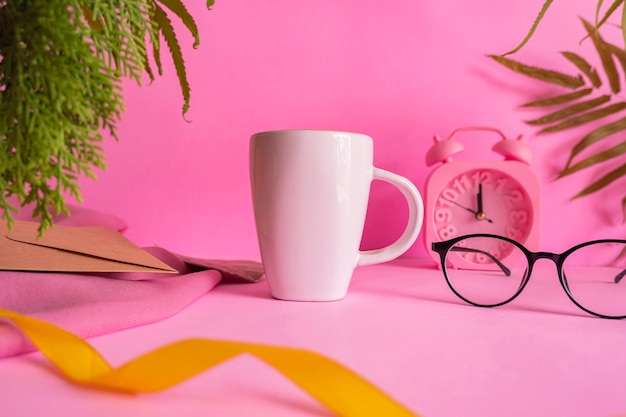 Minimalistische Konzeptidee weiße Kaffeetasse auf rosa Hintergrund mit Weckerdekoration, Blättern und Gläsern