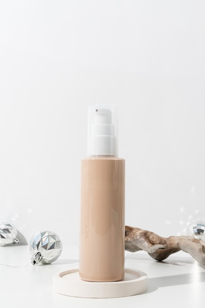 Minimalistische Szene der Foundation-Kosmetikflasche auf dem Podium mit weihnachtlichen Zierkugeln und Kiefer auf hellem Hintergrund