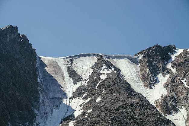 Mínimo paisaje de montaña con una hermosa pared de montaña con glaciar y cornisa de nieve en la parte superior. Paisaje alpino minimalista con grandes montañas nevadas bajo un cielo azul claro. Rocas nevadas iluminadas por la luz del sol.