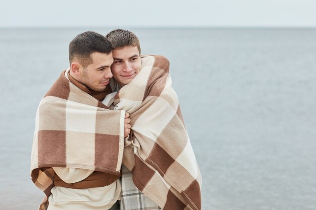 Mínimo retrato de joven pareja gay abrazada en la playa envuelto en una manta con agua en el fondo, espacio de copia