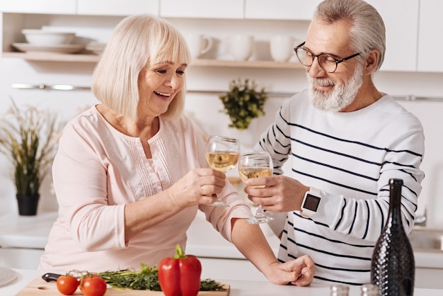 Minuten echten Glücks. Positive glückliche ältere Partner, die in der Küche stehen und Abendessen kochen, Liebe ausdrücken und Champagner trinken