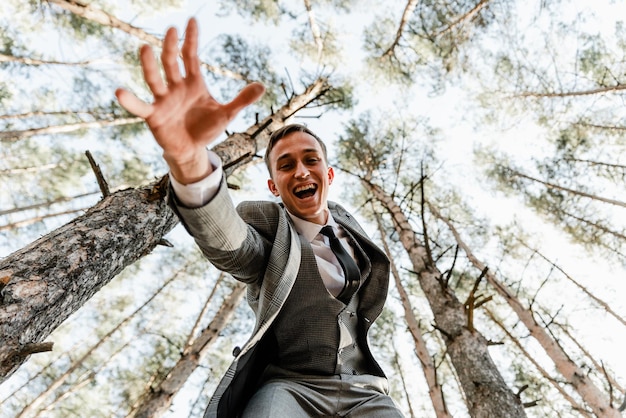 Mira desde abajo al novio de traje que está en el bosque. novio con una pierna levantada. un chico de apariencia europea se está divirtiendo. diferentes emociones de un hombre. novio el día de la boda