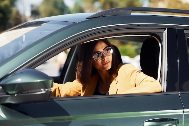 Mira hacia afuera desde el automóvil Joven mujer de moda con abrigo de color burdeos durante el día con su automóvil