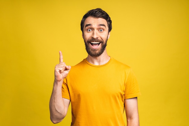 ¡Mira allá! Feliz joven apuesto barbudo en casual apuntando hacia arriba y sonriendo mientras está de pie aislado sobre fondo amarillo.