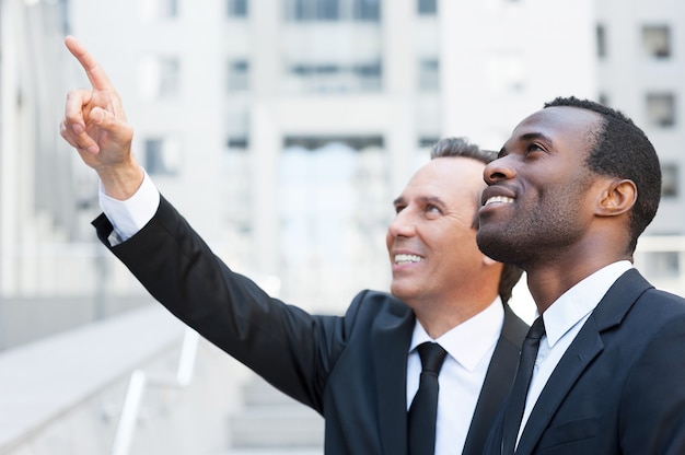 ¡Mira allá! Vista lateral de dos hombres de negocios alegres hablando y gesticulando mientras está de pie al aire libre