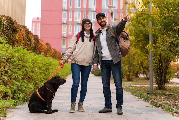 Mira allí. Encantadora pareja pasear junto con el perro al aire libre mientras el hombre apunta hacia afuera con su dedo y sonriendo con dientes