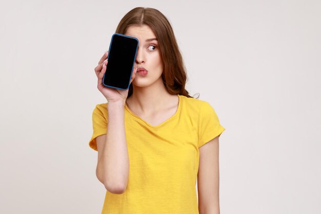 ¡Mira, anúnciate aquí! Retrato de mujer divertida de edad joven en camiseta que cubre la mitad del teléfono de la cara con pantalla en blanco, tiene una expresión de duda reflexiva. Disparo de estudio interior aislado sobre fondo gris.