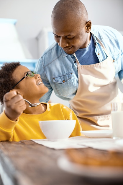 Mira con cariño. Agradable joven en un delantal hablando con su hijo y sonriéndole con cariño mientras come cereales para el desayuno