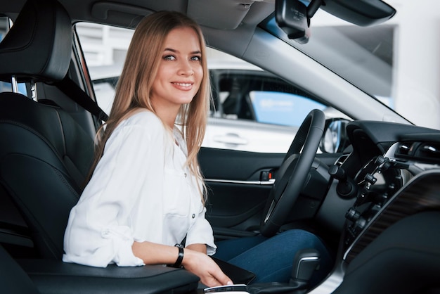 Mira directamente a la cámara una hermosa chica rubia sentada en el coche nuevo con interior negro moderno