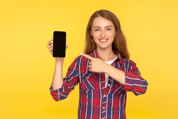 Mira el dispositivo Retrato de una adorable chica guapa con camisa a cuadros apuntando al celular en la mano y sonriendo publicidad de una aplicación móvil o una foto de estudio interior aislada en un fondo amarillo