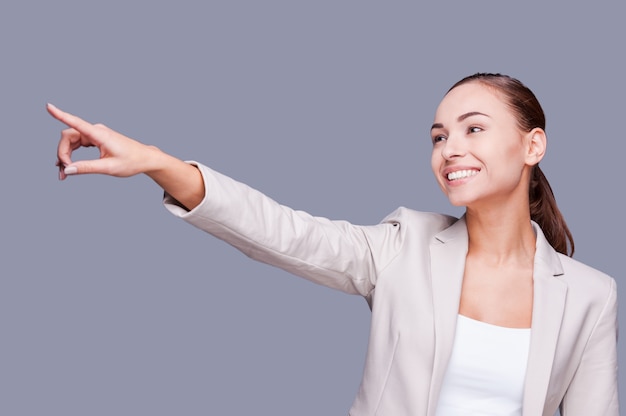 ¡Mira eso! Sonriente joven empresaria apuntando hacia otro lado mientras está de pie contra el fondo gris