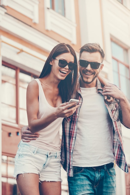 ¡Mira esta imagen! Vista de ángulo bajo de la hermosa joven pareja amorosa de pie juntos al aire libre y mirando el teléfono móvil