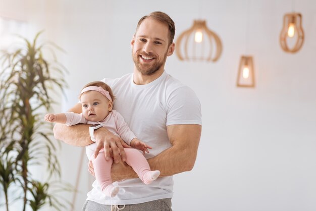 Mira a mi lindo bebé. Sonriente padre barbudo positivo de pie en casa y sosteniendo a su bebé mientras expresa positividad y cuidado