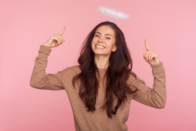 Mira mi nimbo. Retrato de una joven encantadora que señala el halo por encima de la cabeza y mira a la cámara con una sonrisa amable, expresando una belleza angelical. tiro de estudio interior aislado sobre fondo de color rosa