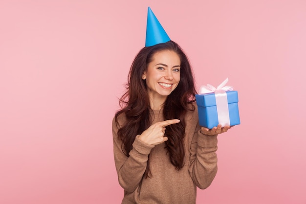 Mira mi presente. Retrato de una joven alegre que usa un cono de fiesta en la cabeza y señala una caja de regalo, sonriendo a la cámara, con una sorpresa de cumpleaños. tiro de estudio interior aislado sobre fondo de color rosa