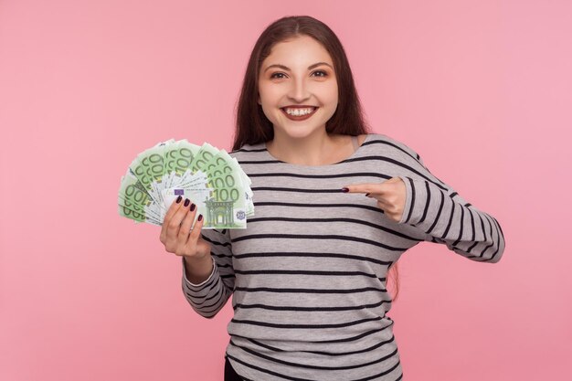 Mira mis ingresos Retrato de una mujer morena feliz con una sudadera a rayas señalando billetes en euros y sonriendo a la cámara satisfecha con la lotería gana un gran premio aislado en un estudio de fondo rosa