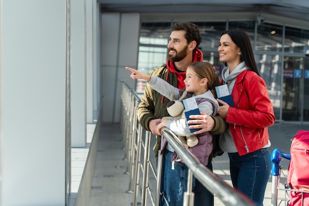 Mira esto. Una niña linda señalando con el dedo algo mientras está de pie cerca de la ventana con sus adorables padres en el aeropuerto. Foto de stock