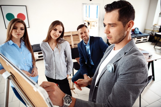 ¡Mira nuestros resultados! Apuesto joven con gafas de pie cerca de la pizarra y apuntando en el gráfico mientras sus compañeros de trabajo escuchan y se sientan en la mesa