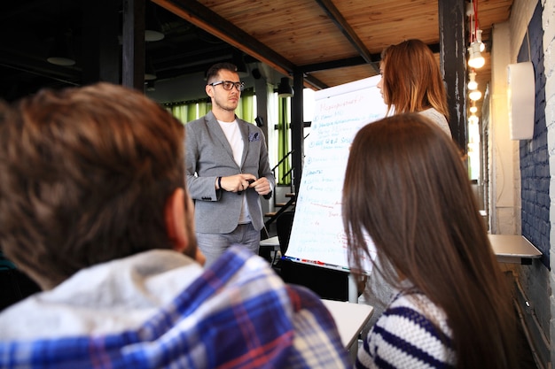 Foto ¡mira nuestros resultados! apuesto joven con gafas de pie cerca de la pizarra y apuntando en el gráfico mientras sus compañeros de trabajo escuchan y se sientan en la mesa