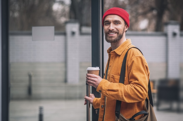 Mirada alegre. Hombre con sombrero rojo y chaqueta naranja con una taza de café en las manos mirando alegre