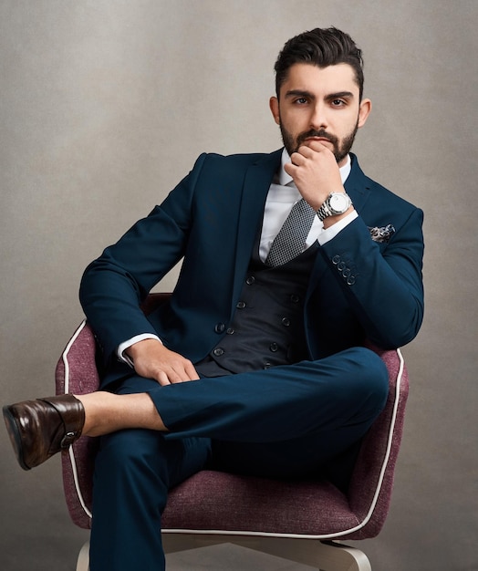 La mirada del éxito Retrato de estudio de un joven hombre de negocios elegantemente vestido sentado en una silla contra un fondo gris