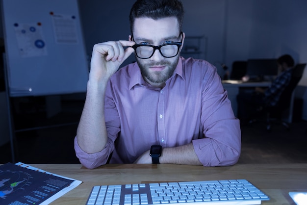 Mirada inteligente. Hombre guapo encantado inteligente sentado frente a la computadora y arreglando sus gafas mientras te mira