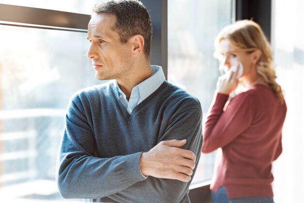 Mirada pensativa. Triste triste infeliz hombre de pie con las manos cruzadas y mirando por la ventana mientras tiene una pelea con su novia