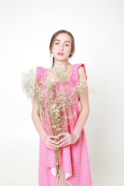 Mirada romántica rizada, ojos hermosos. Flores silvestres blancas en las manos. Chica vestido de luz blanca y cabello rizado, retrato de mujer con flores sobre fondo blanco, pureza e inocencia