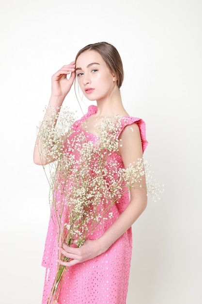 Mirada romántica rizada, ojos hermosos. Flores silvestres blancas en las manos. Chica vestido de luz blanca y cabello rizado, retrato de mujer con flores sobre fondo blanco, pureza e inocencia