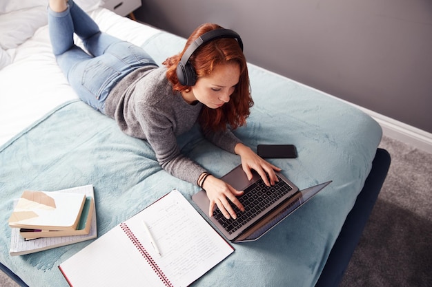 Mirando hacia abajo a una estudiante universitaria usando audífonos acostado en la cama trabajando en una computadora portátil