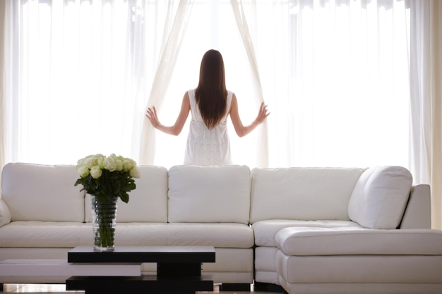 Mirando el amanecer Una mujer joven mirando a través de la ventana de su sala de estar temprano en la mañana