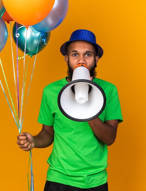 Mirando a la cámara joven afroamericano con gorro de fiesta sosteniendo globos habla por altavoz