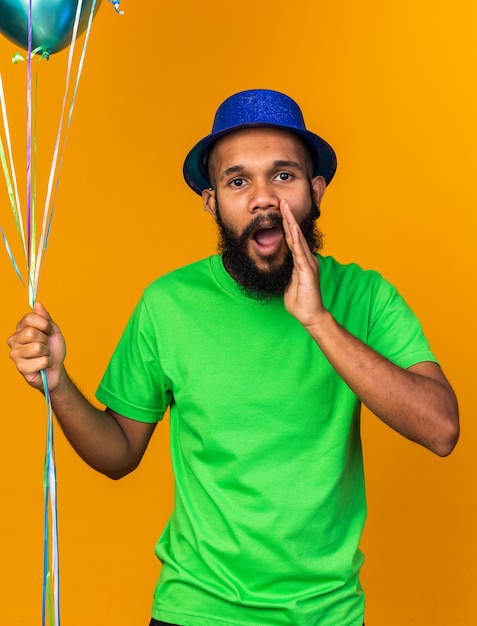 Mirando a la cámara joven afroamericano vistiendo gorro de fiesta sosteniendo globos llamando a alguien