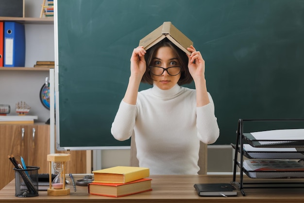 mirando a la cámara una joven maestra con gafas poniendo un libro en la cabeza sentada en un escritorio con herramientas escolares en el aula