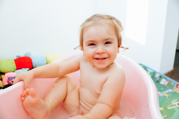 Mirando a la cámara, una niña sentada en el baño, levanta una pierna, sostiene la bañera, sonríe, ríe.